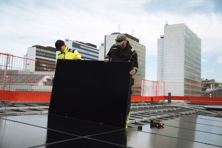 Two workers installing a solar panel on the roof of a building in Stockholm, with city skyscrapers in the background.