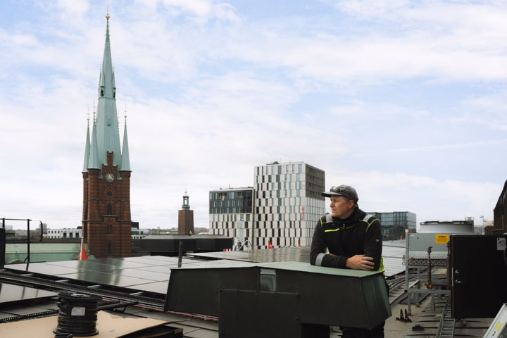 Man in work clothes looking out over Stockholm rooftop with solar panels and cityscape in background.